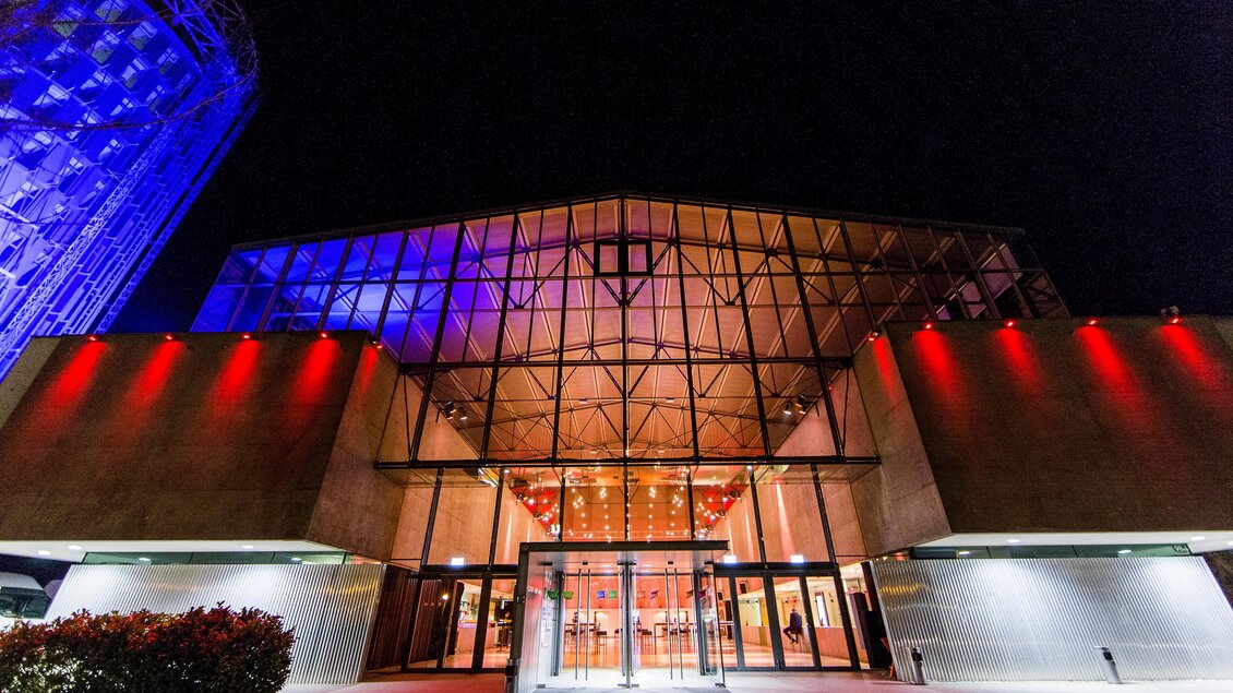 Ein modernes Gebäude mit großen Glasfenstern, beleuchtet in roten und blauen Lichtern. Der Eingangsbereich strahlt eine einladende Atmosphäre aus. | © -hlh_photoworkers