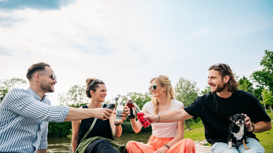Vier Freunde stoßen mit Getränken am Fluß an. | © Graz Tourismus - Mias Photoart