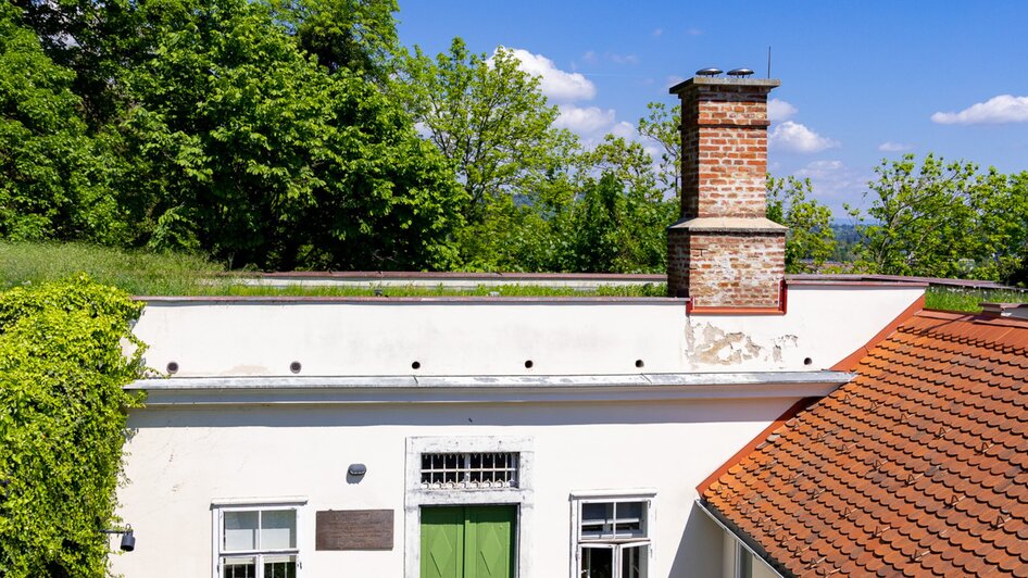 Das Cerrini-Schlössl mit grünem Eingang und Kamin, umgeben von Bäumen und blauem Himmel.