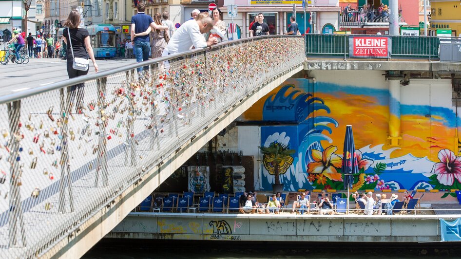 Überblick über die Mur und die bunten Graffitis in Graz, mit einer Brücke voller Liebesschlösser.