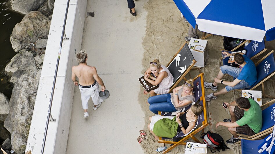 Menschen genießen die Sonne am Murufer, während andere vorbeigehen.