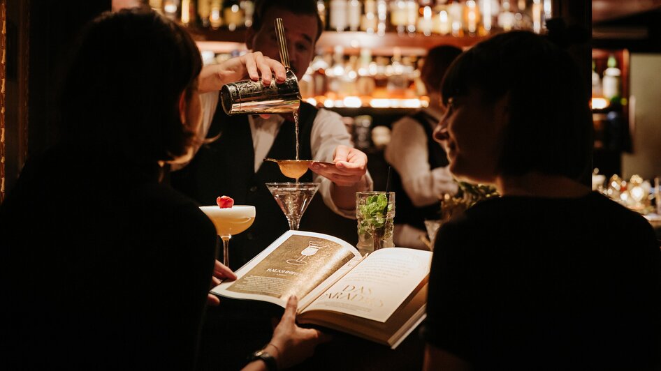 Zwei Gäste in der Bar, während ein Barkeeper Cocktails zubereitet.