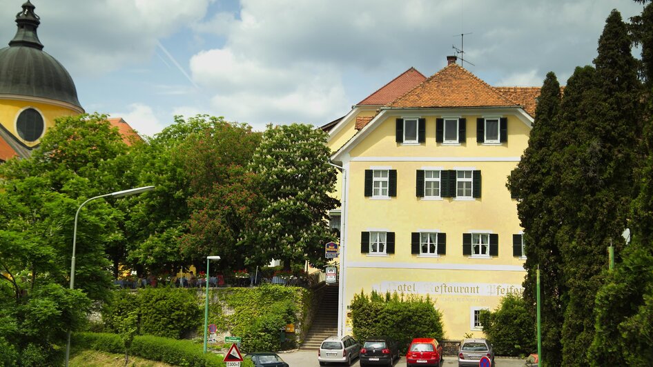Das Gebäude des Kirchenwirt Mariatrost in Graz, umgeben von Bäumen und Autos.
