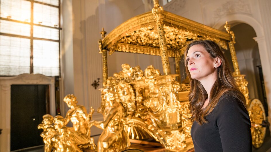 Eine Frau steht vor einem kunstvoll verzierten goldenen Prunkwagen.