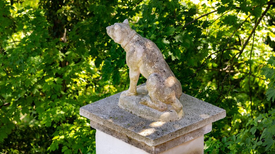 Eine Steinskulptur eines Hundes auf einem Podest, umgeben von grünen Blättern. | © Graz Tourismus - Harry Schiffer