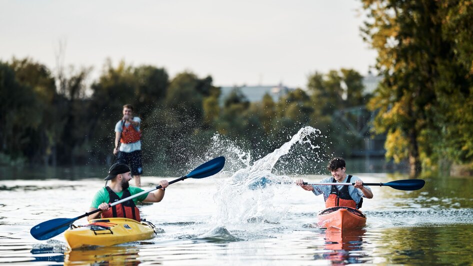 Zwei Kajakfahrer paddeln auf ruhigem Wasser, während ein Dritter auf einem SUP steht und zuschaut.