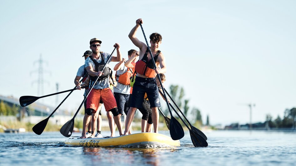 Gruppe von Menschen paddelt auf einem großen SUP im Wasser.