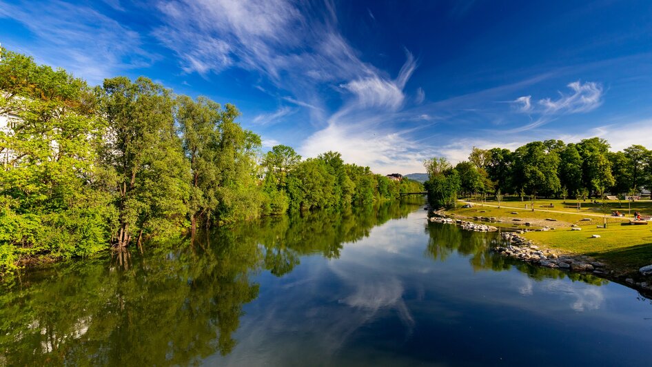 Idyllische Szenerie an der Augartenbucht in Graz mit spiegelndem Wasser und grünem Ufer.