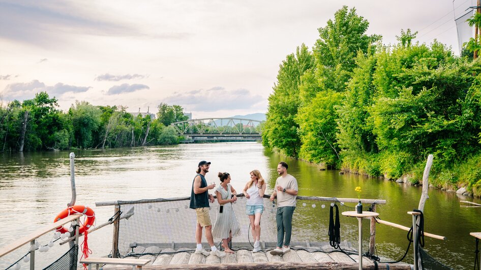 Gruppe von Freunden genießt Zeit am Fluss in Graz, umgeben von Bäumen.